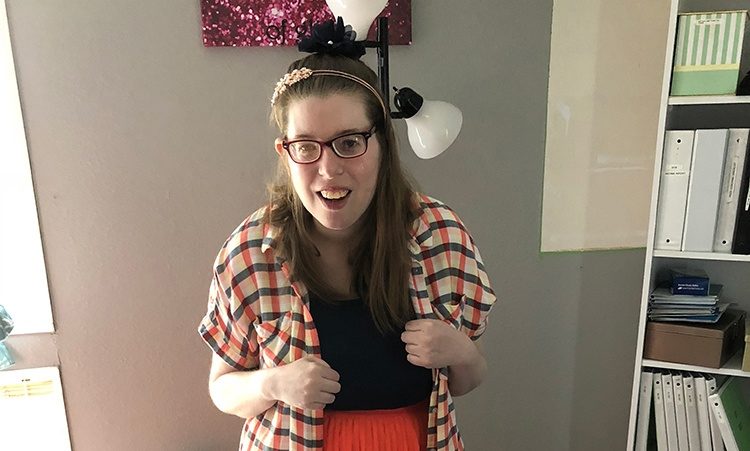 A young woman wearing a plaid shirt, wearing glasses, smiling at camera. Indoor setting.
