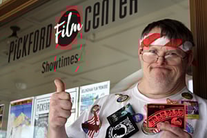 A man wearing glasses holding a free pass for a film, standing in front of the movie theater, thumb up