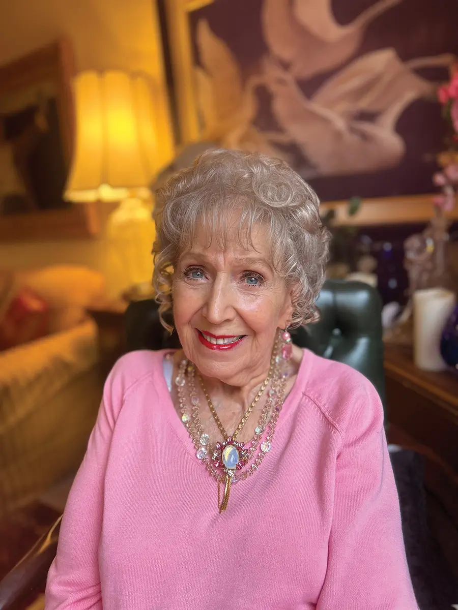 Betty wearing a pink long sleeve shirt with beautiful necklesses, smiling.