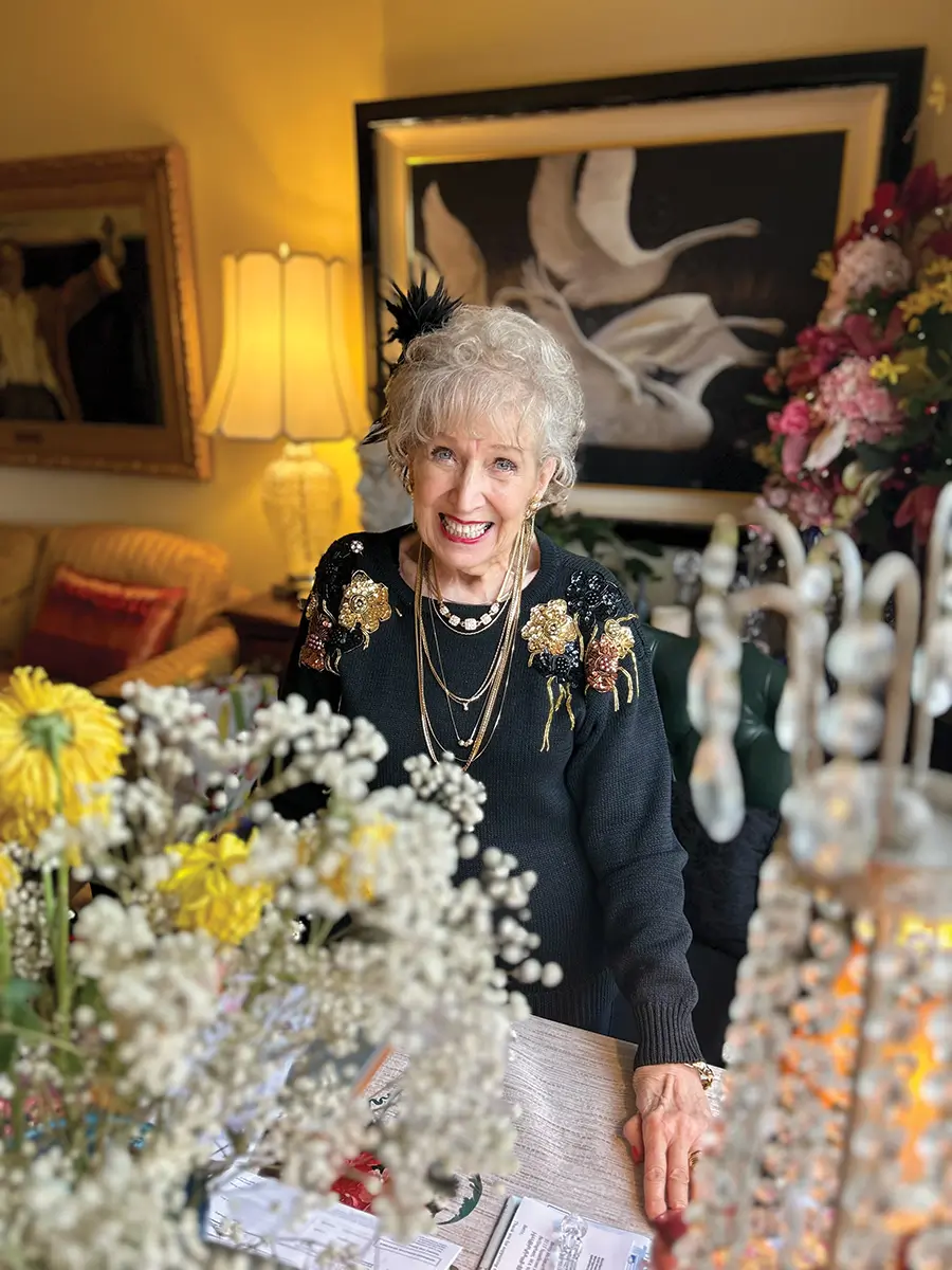 Betty wearing black long sleeve shirt. flowers in the front, and picture frames on the wall in the background.