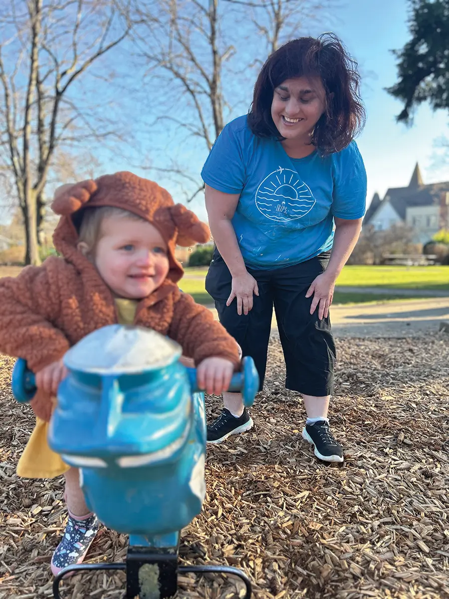 Hilary wearing a light blue t-shirt and a black pants, slightly bending over, smiling, watching a child at the park