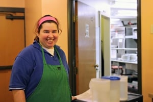 A woman waring a blue short sleeve shirt, a green apron, smiling, at work environment
